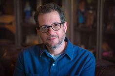 a man wearing glasses and a blue shirt looks at the camera while sitting in front of a bookcase