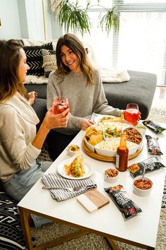 two women sitting at a table with food and drinks in front of them on the coffee table