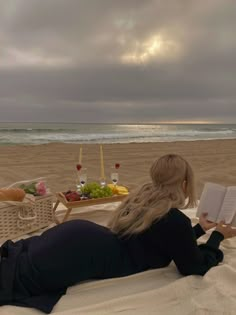 a woman laying on top of a sandy beach next to the ocean reading a book