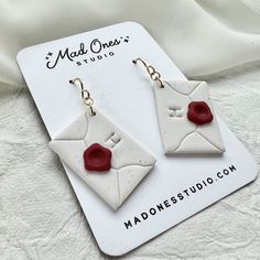 a pair of white and red ceramic earrings on top of a card board with the logo mad ones studio