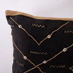 a black pillow with gold thread and buttons on the front, sitting against a white wall