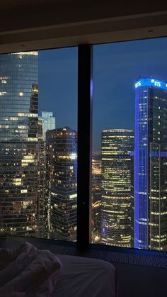 a view of the city at night from a hotel room with a large window that looks out onto skyscrapers