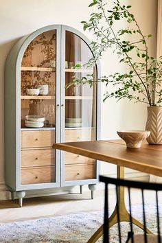 a wooden table topped with a vase filled with green plants next to a glass cabinet