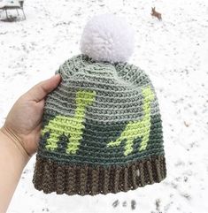 a hand holding up a crocheted hat with a green and yellow dinosaur on it