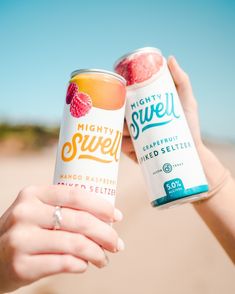 two people holding up cans of fruity sweet drink on the beach, one has a strawberry in it