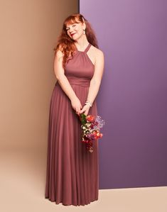 a woman in a long brown dress holding a bouquet and standing next to a purple wall