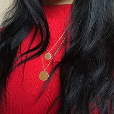 a woman with long black hair wearing a red sweater and two gold disc necklaces