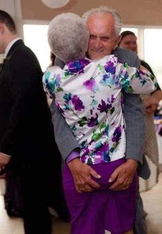 an older man and woman hugging each other in a room full of people standing around