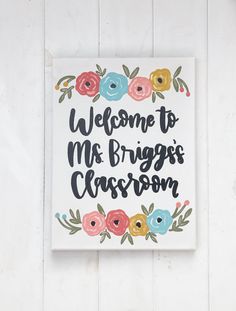 a welcome sign hanging on the side of a white wall with flowers and leaves around it