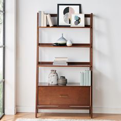 a shelf with books, vases and pictures on it next to a window in a white room