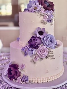 a three tiered wedding cake with purple flowers on the side and pearls around the edges