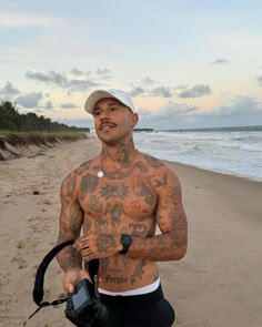 a man with tattoos on his chest holding a camera in front of the ocean and sand