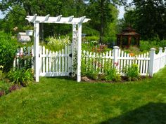 a white garden gate surrounded by lush green grass and flowers in the foreground, with a gazebo in the background