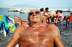 an older man with tattoos on his chest sitting in a chair near the water and other people