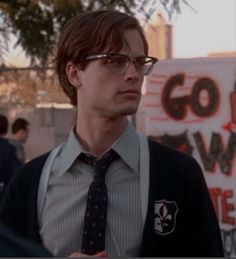 a man wearing glasses and a tie standing in front of a sign with writing on it