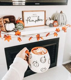 a person holding a mug in front of a fireplace with fall decorations on the mantle