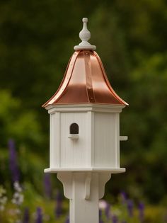 a white bird house with a copper roof in front of some purple and white flowers