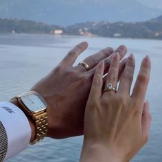 two people holding their hands up to each other near the water with mountains in the background