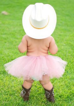 a baby girl wearing a cowboy hat and pink tutu skirt, standing in the grass