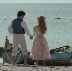 a man and woman holding hands while walking on the beach with boats in the background