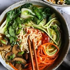 two bowls filled with noodles and vegetables next to chopsticks on a white table