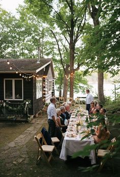 a group of people sitting around a table in front of a house with lights strung from the trees