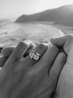 a man and woman holding each other's hand near the ocean with an engagement ring on their finger