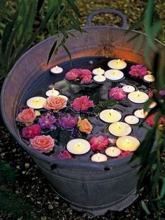 candles floating in a bucket filled with water surrounded by pink and red flowers on the ground