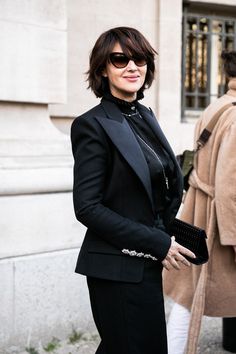 a woman in a black suit and sunglasses is walking down the street with her hand on her hip