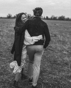 a man and woman walking in an open field holding each other's back as they look at the sky