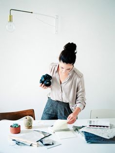 a woman standing at a table holding a camera in one hand and looking at papers on the other