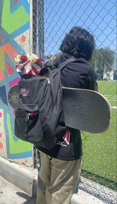 a person with a skateboard in their back pack standing by a fence and looking at graffiti on the wall