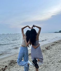 two young women standing on the beach making heart shapes with their hands and looking at the ocean