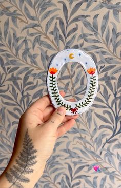 a hand holding a paper wreath with flowers and leaves on the inside, in front of a wallpapered background