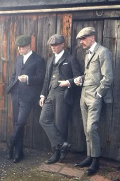 three men in suits and hats standing next to a wooden wall with one man looking at his phone