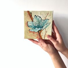 a hand holding up a piece of art with a blue flower on it's side