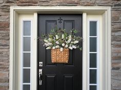 a black front door with a basket full of flowers on the side and a brick wall behind it