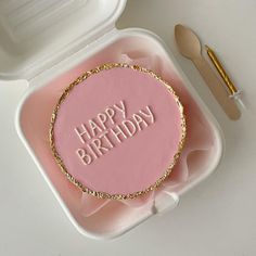 a pink birthday cake with the words happy birthday on it in a plastic container next to utensils