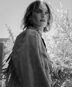 a woman standing in front of a tree wearing a fringed jacket and looking off to the side