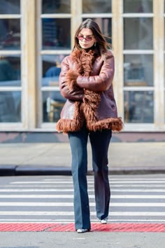 a woman walking across a cross walk wearing a fur coat and dark blue pants with her arms crossed