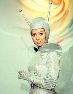 a woman in a silver outfit with two antennae on her head and hands out to the side