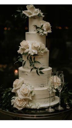 a three tiered cake with white flowers and greenery on the top is surrounded by wine glasses