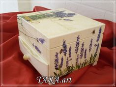 a white box with lavender flowers painted on the front and sides sitting on a red cloth