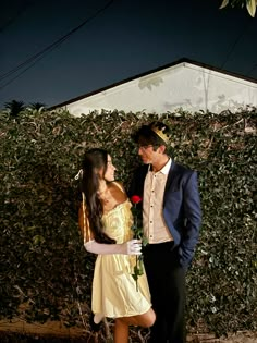 a man and woman standing next to each other in front of a hedge at night