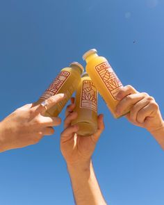 two hands holding three bottles of orange juice in front of a blue sky with white clouds