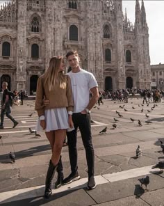 a man and woman standing in front of a large building with pigeons flying around them