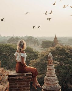 a woman sitting on top of a brick wall next to birds flying over the trees