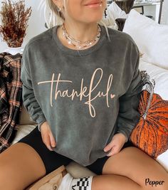 Embrace the cozy vibes of the fall season with our Thankful Sweatshirt, perfect for Thanksgiving and Friendsgiving celebrations! This cute fall sweatshirt features the word "thankful" in a beautiful cursive font, accented with a dainty heart, adding a touch of elegance to your autumn wardrobe. Crafted on a Comfort Colors fall crewneck, this Thanksgiving crewneck sweatshirt is the ideal choice for those chilly days spent at the pumpkin patch or gathering with loved ones. Whether you're dressing u Comfy Fall Tops, Comfy Tops For Fall, Gray Cozy Fit Top For Fall, Cozy Fit Gray Top For Fall, Cozy Fit Crew Neck Sweatshirt For Fall, Comfy Relaxed Fit Tops For Fall, Gray Sweater With Graphic Print For Fall, Comfy Fall Loungewear Sweatshirt, Comfy Cozy Fit Fall Tops