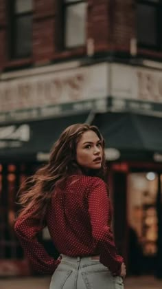 a woman with long hair standing in front of a building wearing a red shirt and grey pants