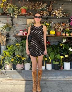 a woman standing in front of potted plants and wearing brown boots with her hands on her hips